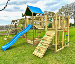 Großer Holzspielturm mit Rutsche, Kletterwand, Wackelbrücke und Schaukeln auf einem Rasen